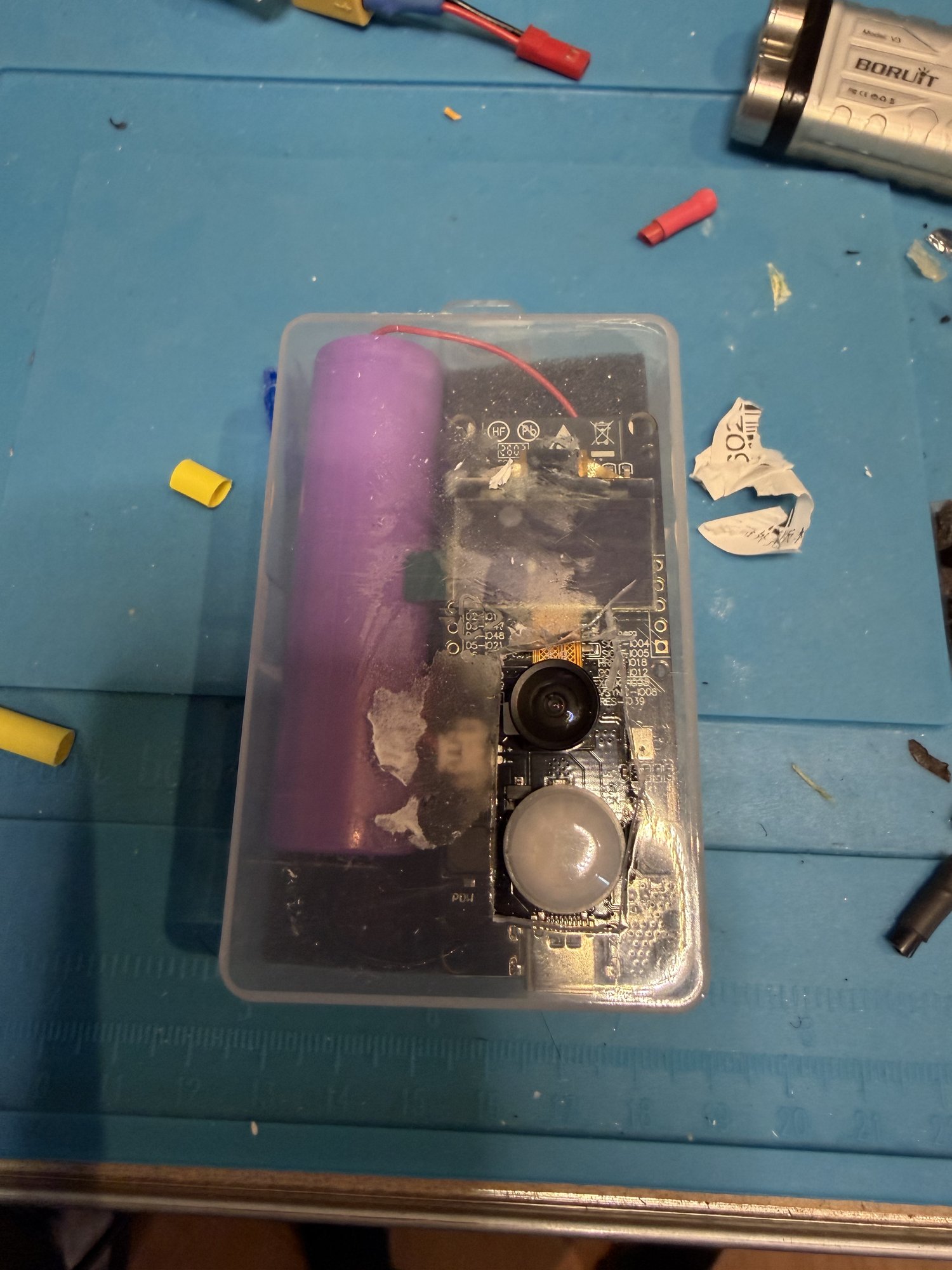 The T-Camera S3 and an 18650 cell in a clear plastic box on the workbench