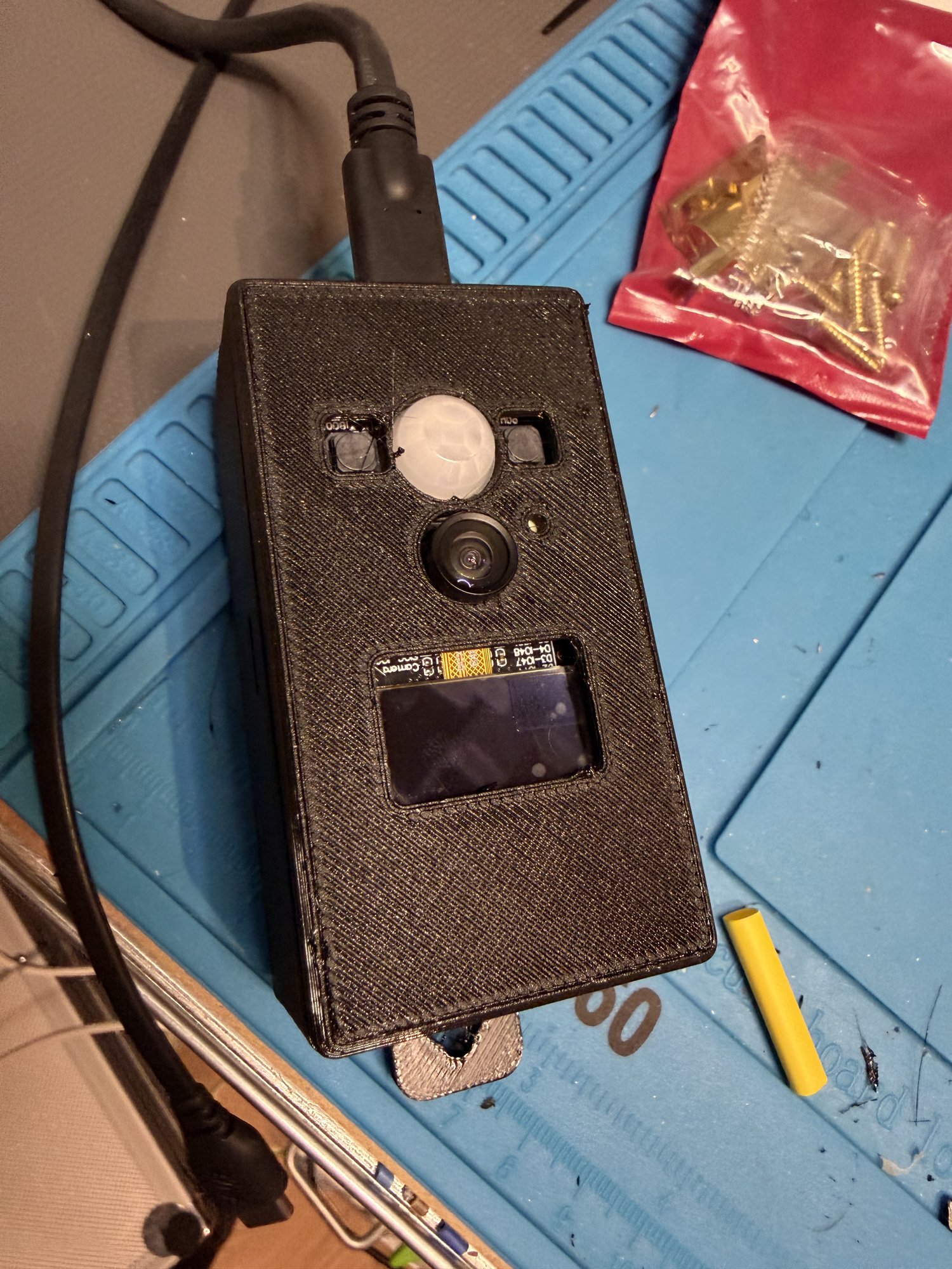 The black 3D-printed case with the PIR dome, camera lens and OLED screen showing through